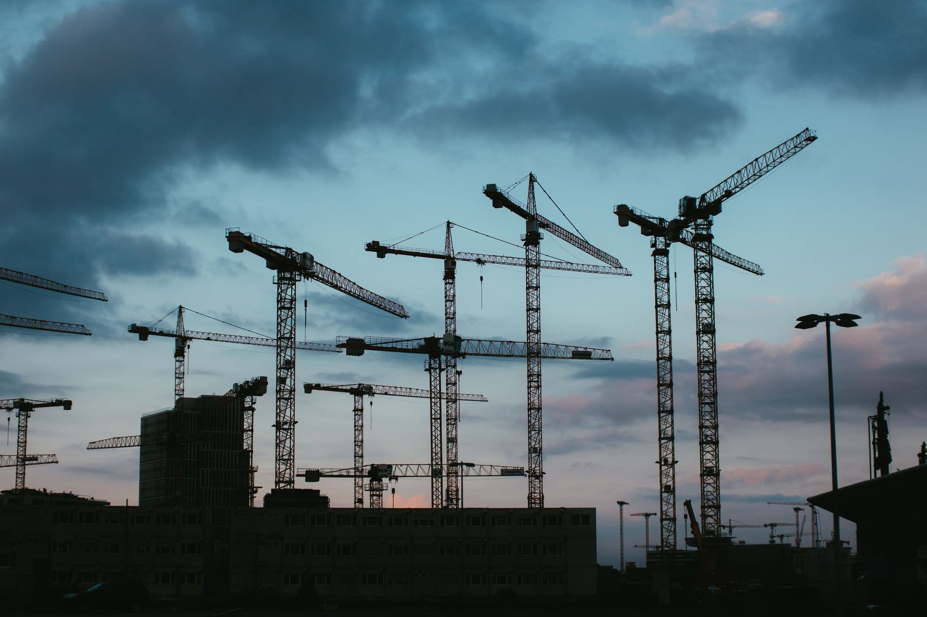 Silhouetten van veel torenkranen boven gebouwen, tegen een bewolkte lucht met een roze gloed