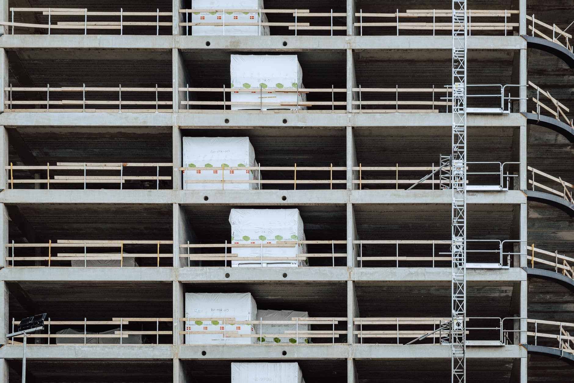 Betonnen verdiepingen in aanbouw met witte ingepakte pakketten, leuningen en een bouwliftmast