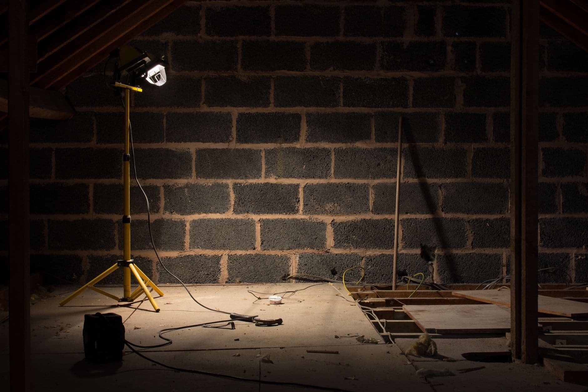 Zolder met een muur van grijze betonblokken, een werflamp op een geel statief en kabels