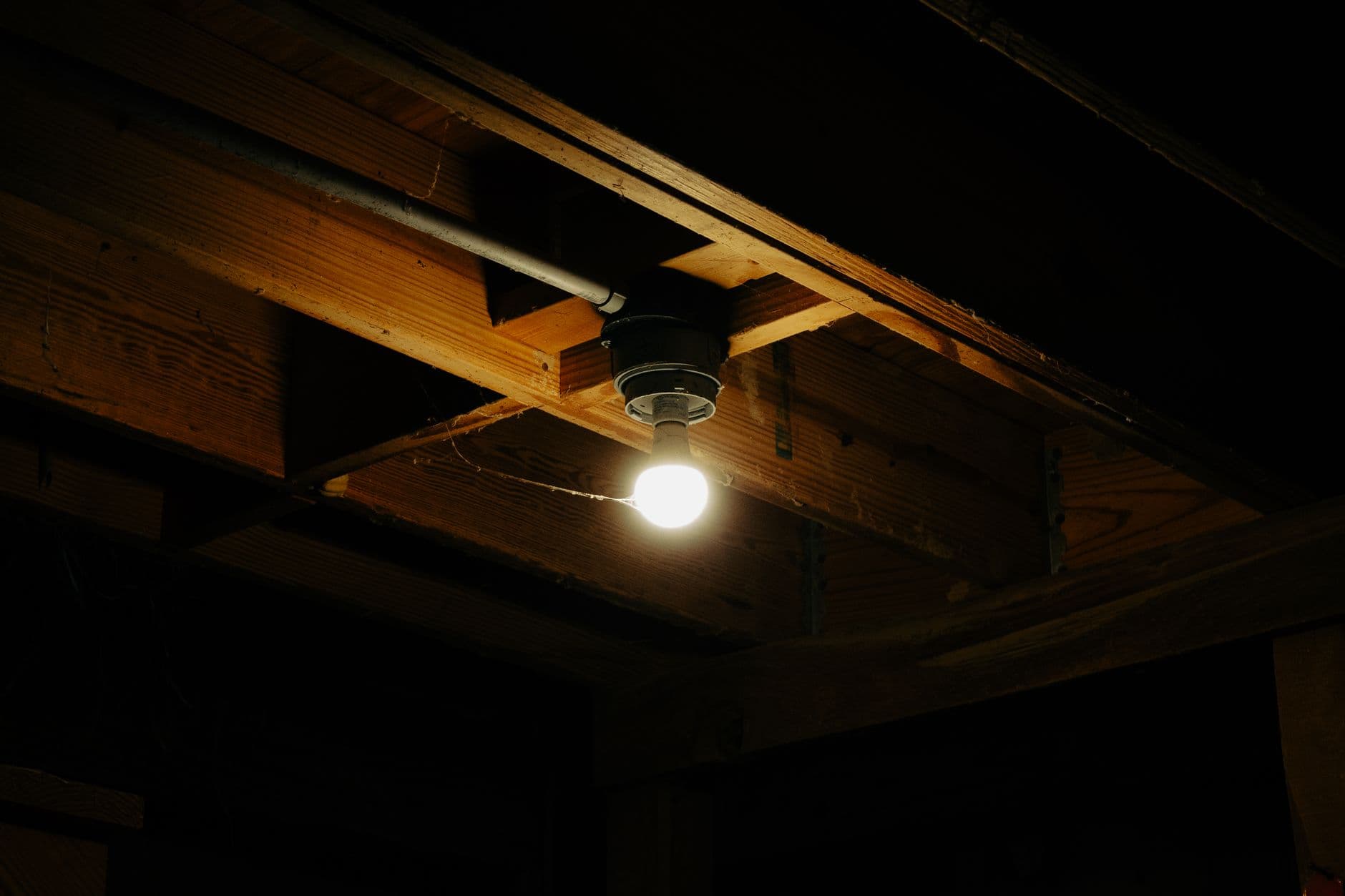 Lamp zonder kap brandt tussen houten balken aan een donker plafond, naast een metalen buis