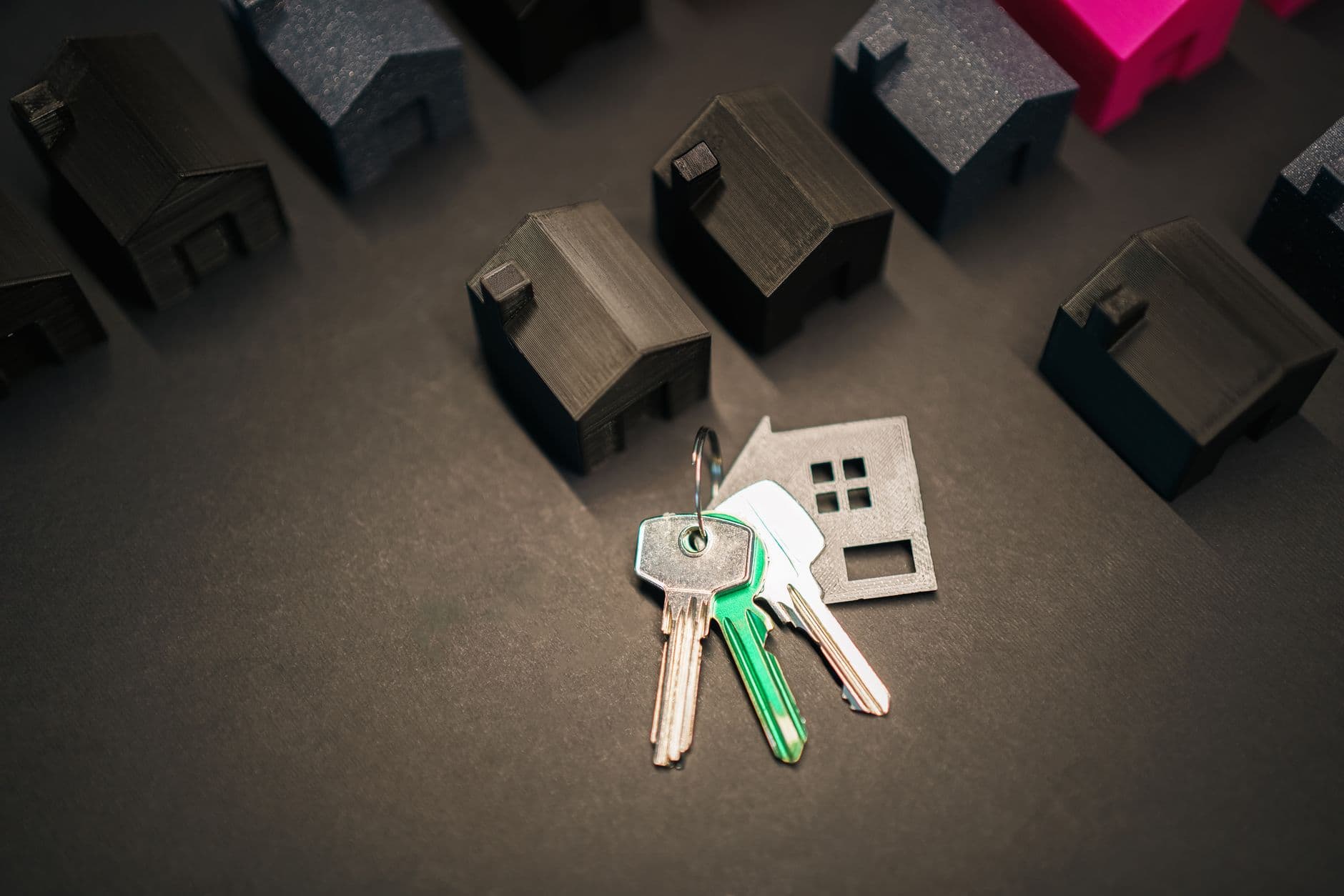 Drie sleutels met een grijze sleutelhanger in de vorm van een huisje, tussen donkere miniatuurhuisjes