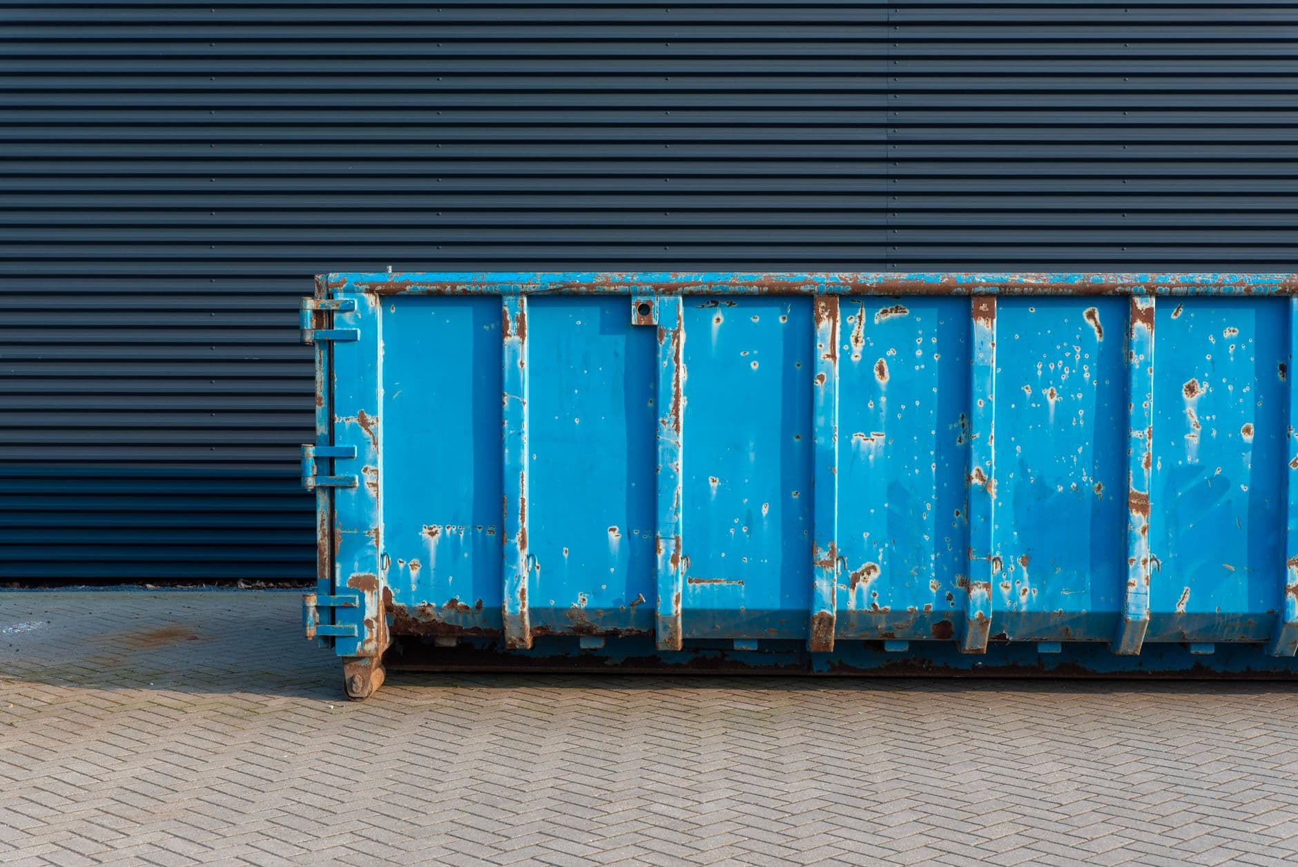 Blauwe afzetcontainer met roestplekken op een klinkerverharding voor een donkere metalen gevel