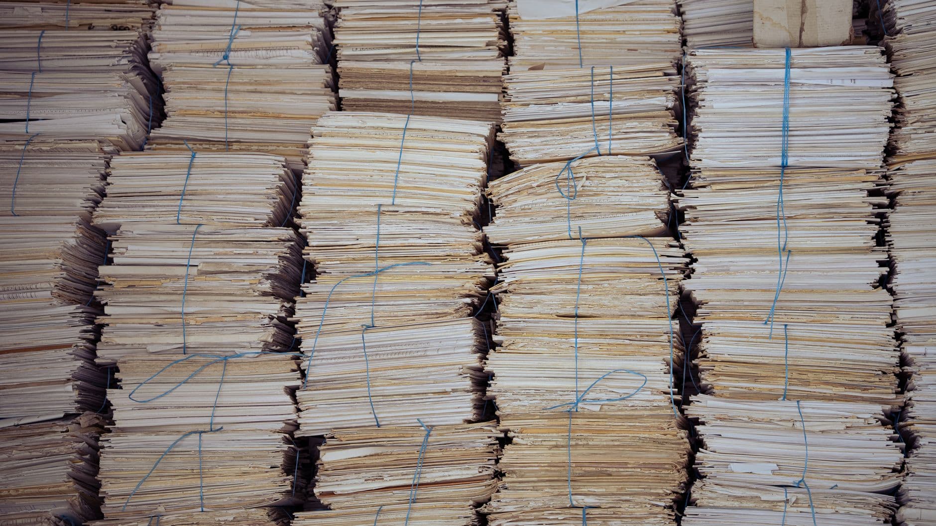 Bundels vergeeld papier met blauwe touwtjes samengebonden, in hoge stapels naast elkaar