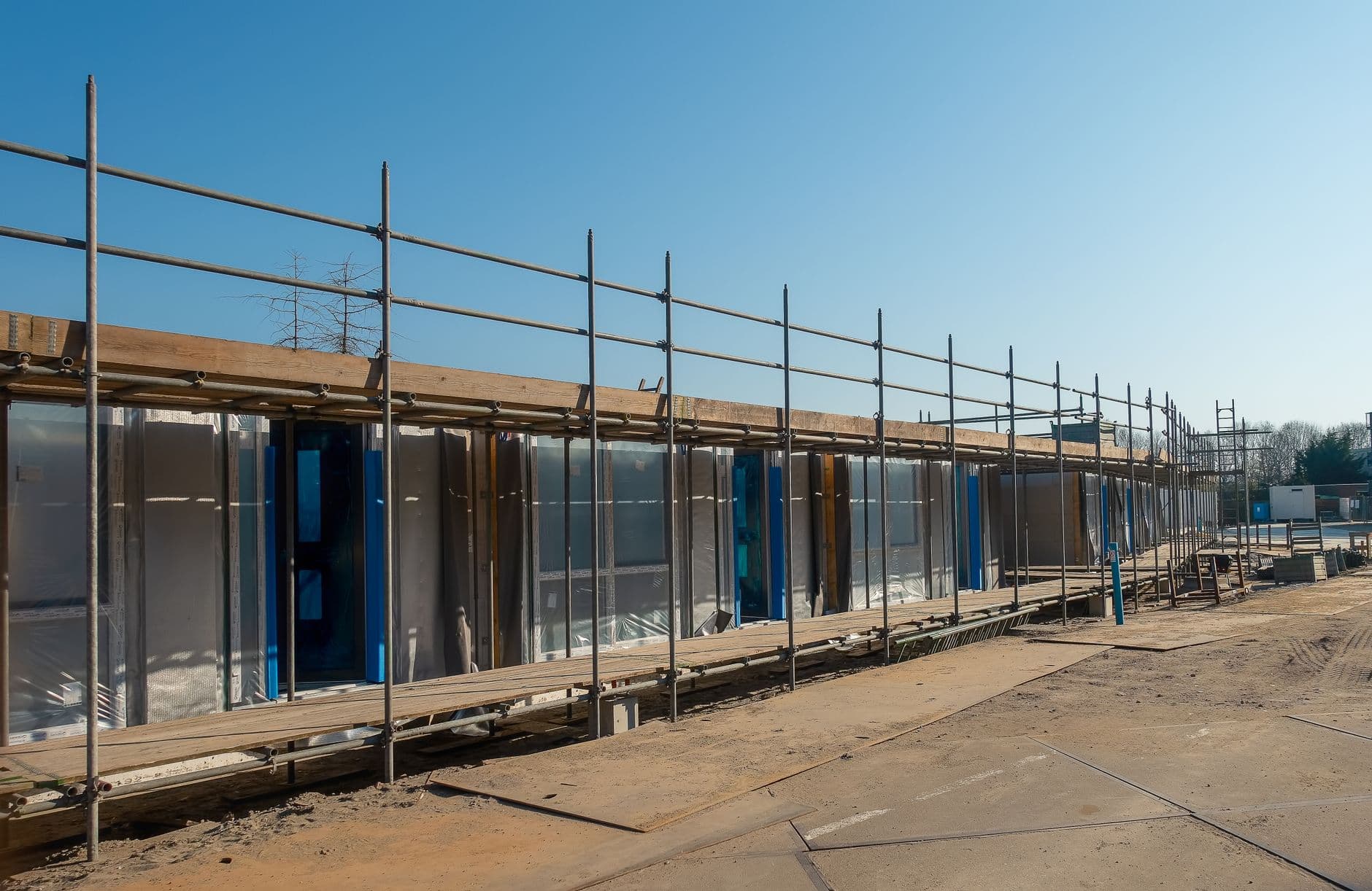 Steiger langs een laag gebouw met plasticfolie over de gevel, onder een blauwe lucht