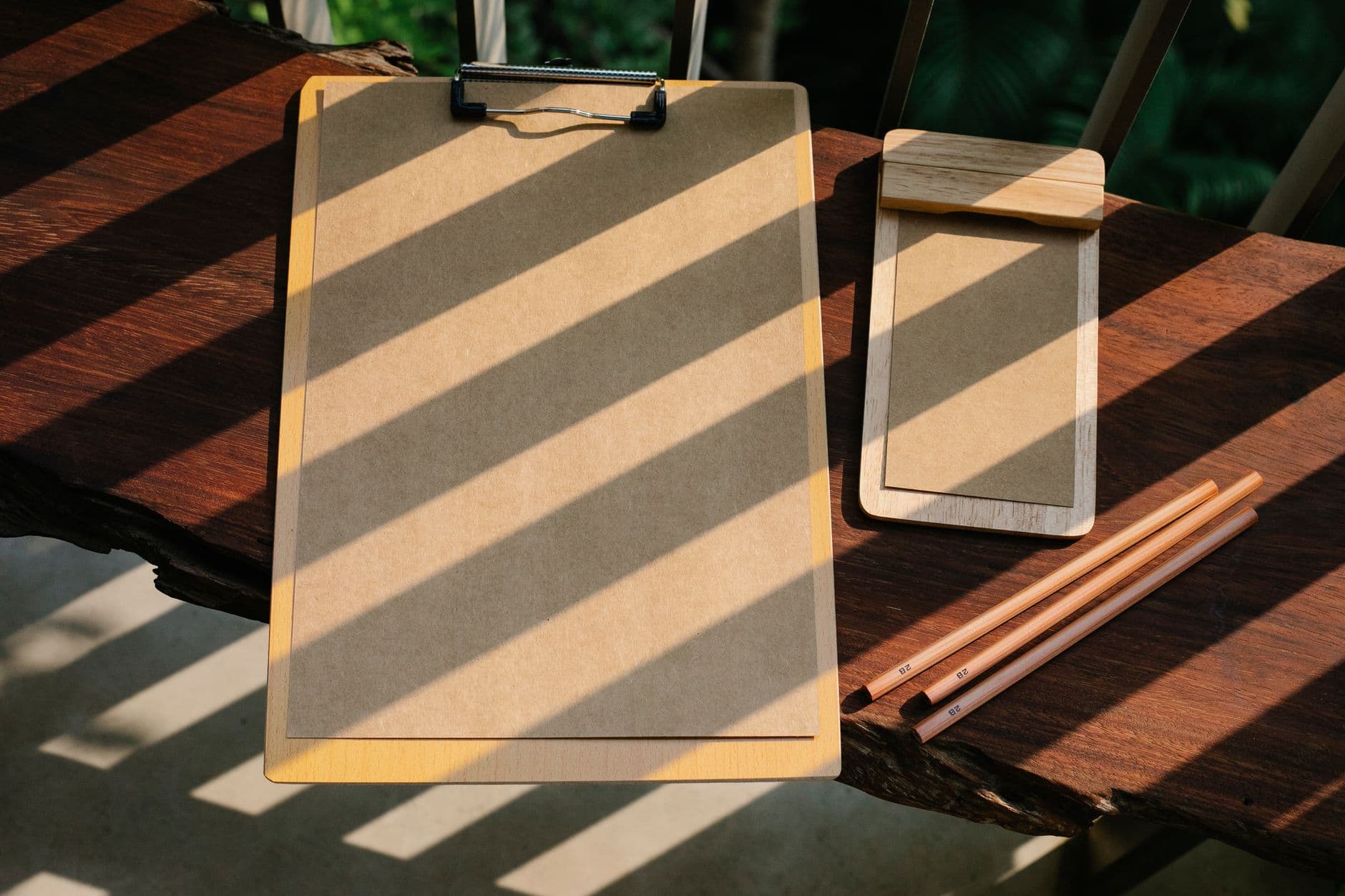 Twee klemborden met bruin papier en drie potloden op een houten tafel met schuine lichtstrepen