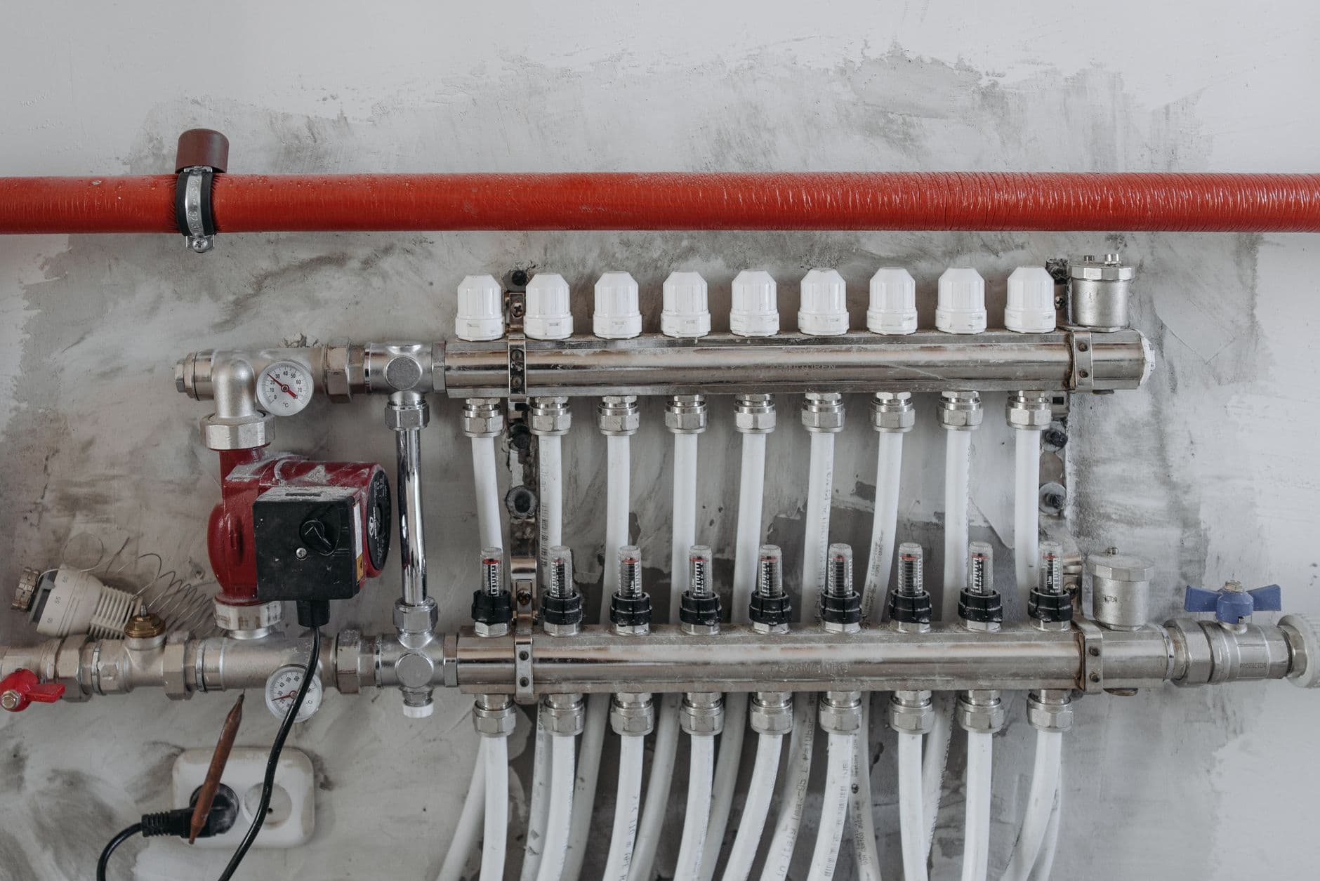 Metalen verdeler met manometer, pomp en een rij witte buizen onder een rode leiding